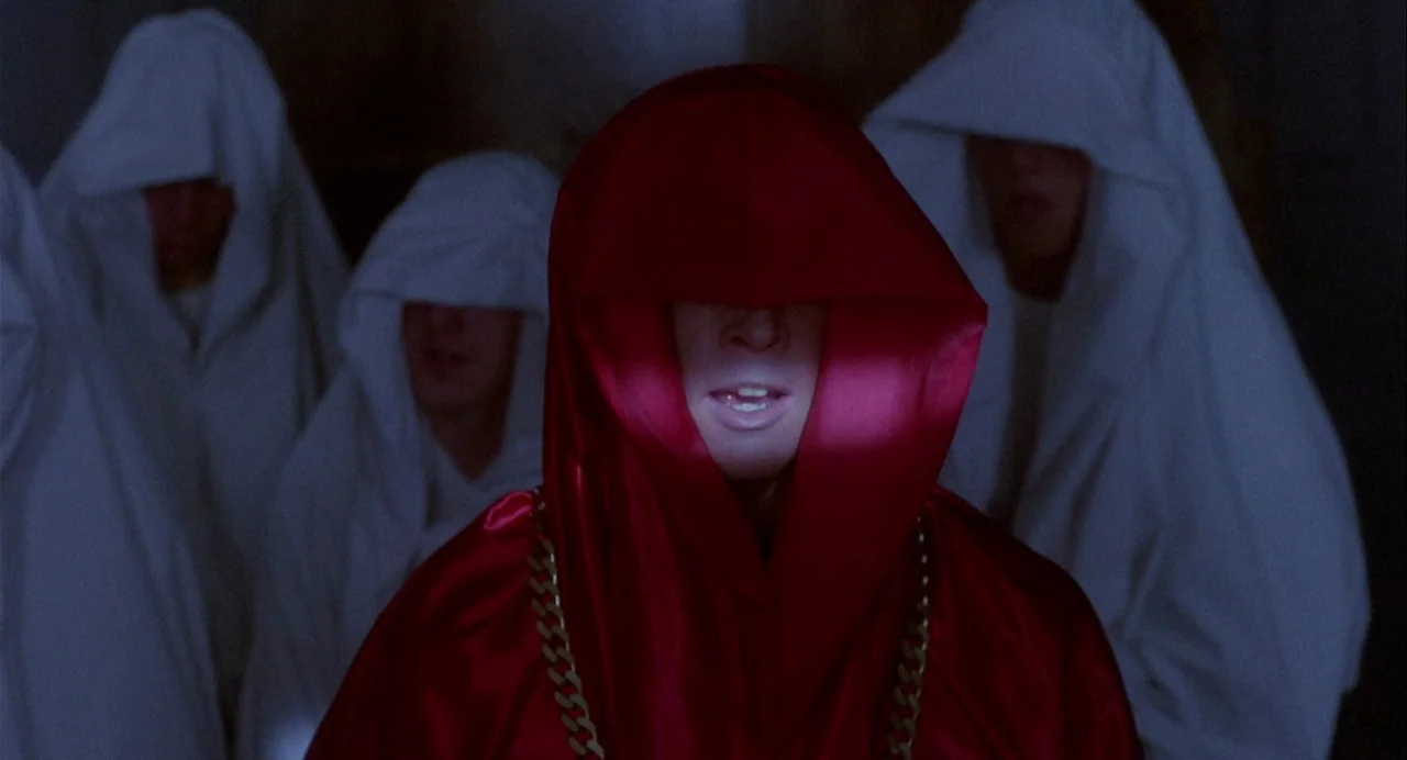 Vamp (1986) movie still: medium — A dark space filled with figures wearing white robes in the background. A central figure wearing a bright red…
