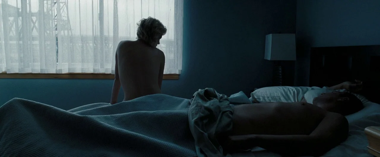 The Burning Plain (2008) movie still: wide — A dimly lit room features a bed positioned beneath a window covered by sheer curtains. The woman sits…