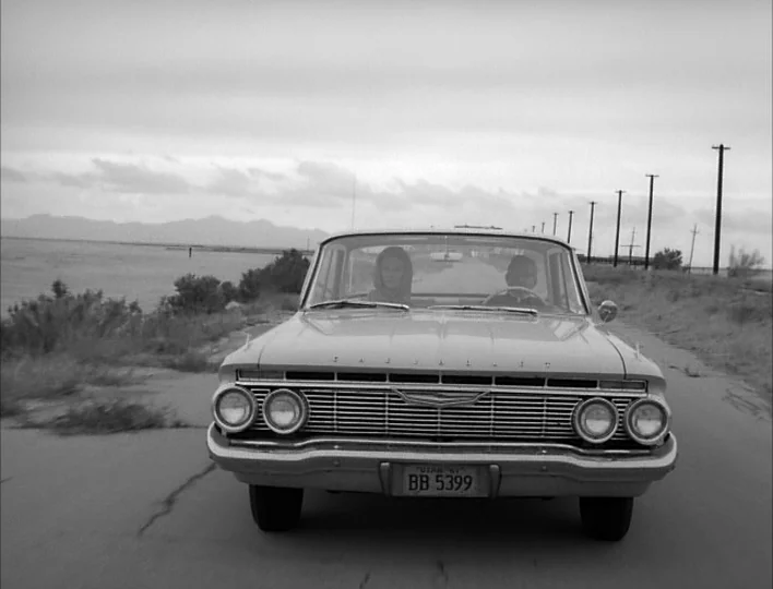 Carnival of Souls (1962) movie still: medium — A vintage 1960s car travels down an empty, desolate road bordered by utility poles. A woman and man…