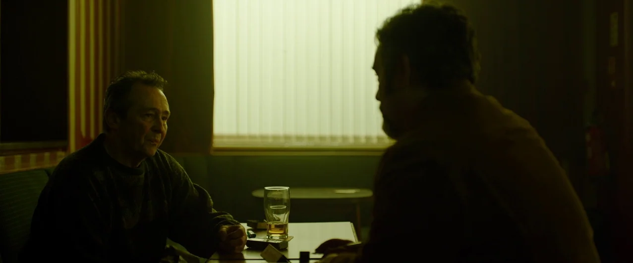 Ghost Stories (2017) movie still: medium — A dimly lit room features a table set with a glass of beer. Two men sit opposite one another in front of…
