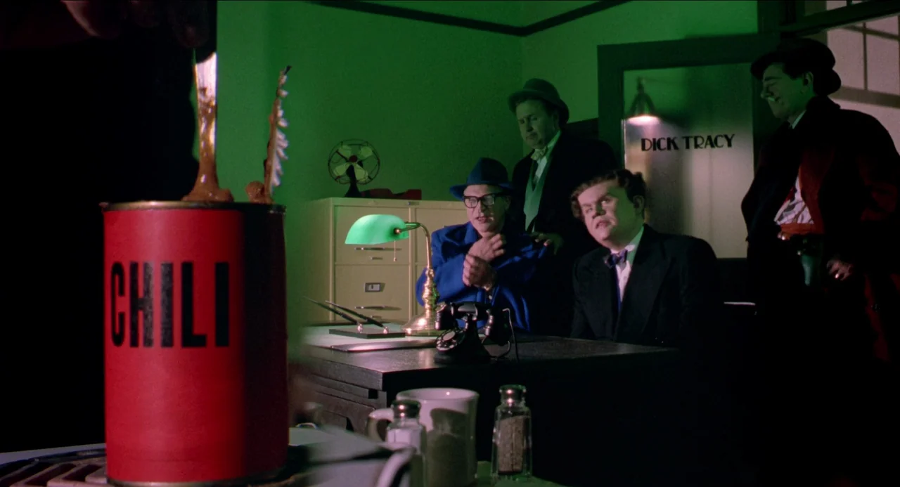 Dick Tracy (1990) movie still: medium — A dimly lit room with green walls contains a desk with men gathered around. A large red can of chili sits in…