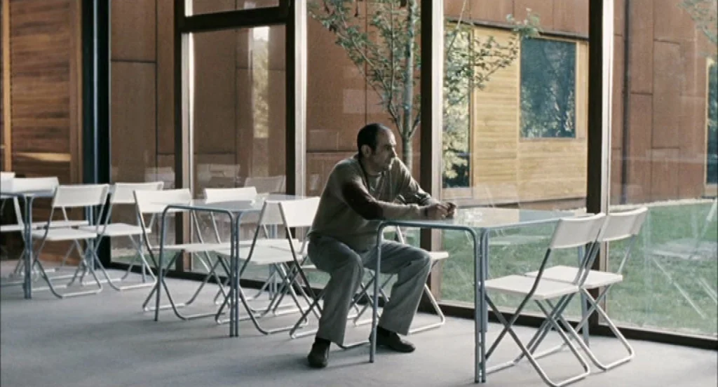 Timecrimes (2007) movie still: medium — A modern cafeteria area with floor-to-ceiling windows showing a green lawn outside. A middle-aged man sits…