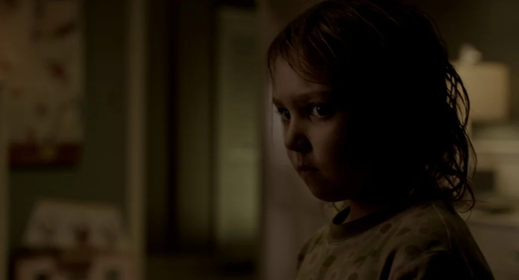 Mama (2013) movie still: closeup — A dimly lit room with blurred interior background details visible in the deep shadows. A young child with messy…