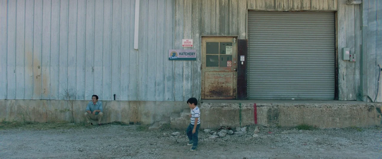 Minari (2020) movie still: wide — A dilapidated industrial hatchery building with a corrugated metal facade and gravel foreground. An adult man…