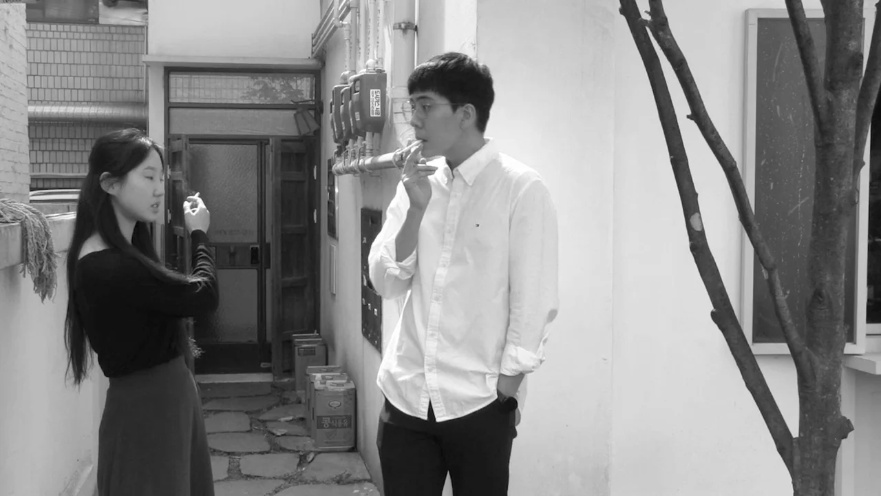 Walk Up (2022) movie still: medium — A stone-paved alleyway features a doorway and utility meters on the white wall. A young man and woman face each…