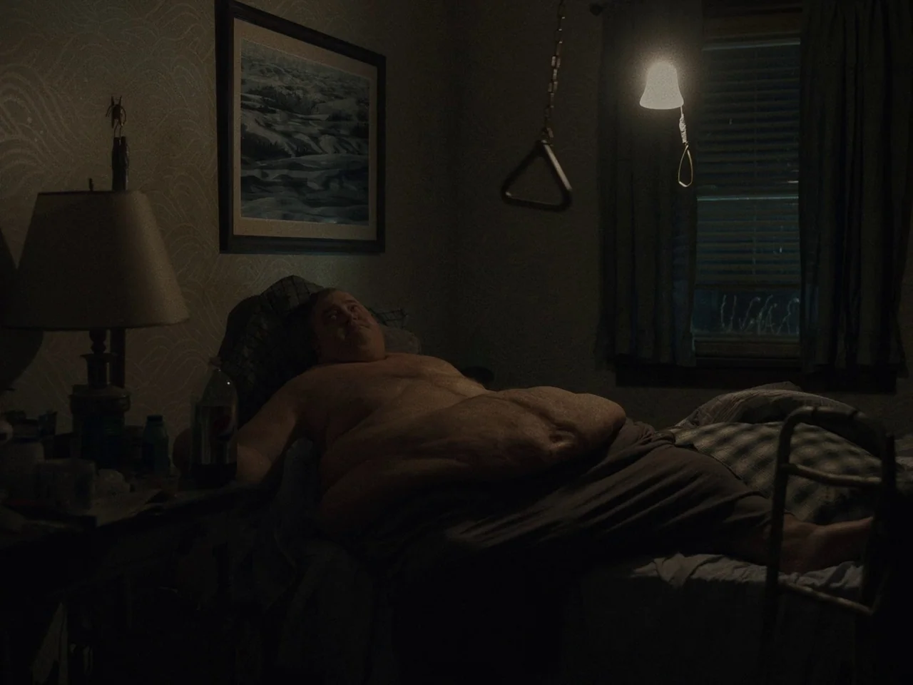 The Whale (2022) movie still: medium — A dark, cluttered bedroom features wallpapered walls and a metal mobility aid hanging overhead. An obese man…