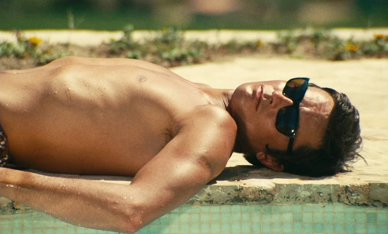 The Swimming Pool (1969) movie still: closeup — A man lies shirtless on the stone perimeter of a swimming pool. Sunlight illuminates his upper body…
