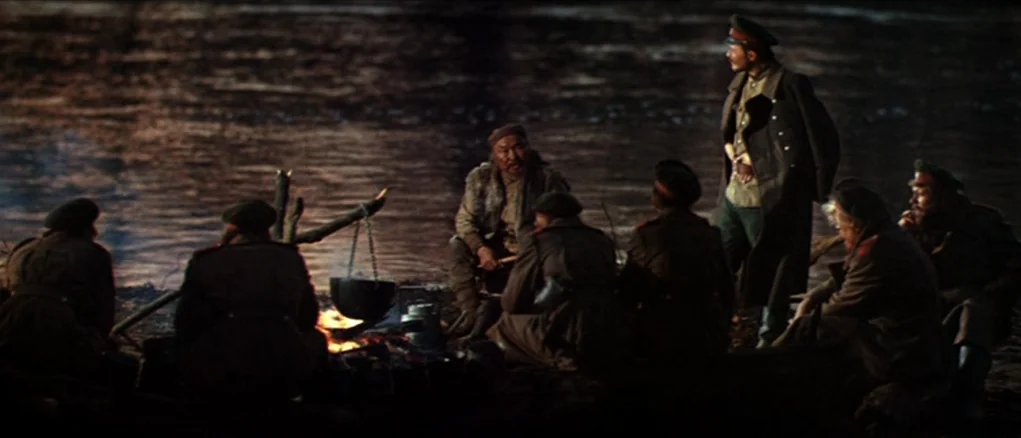 Dersu Uzala (1975) movie still: wide — A group of soldiers gathers around a small campfire beside a dark river at night. One officer stands tall…