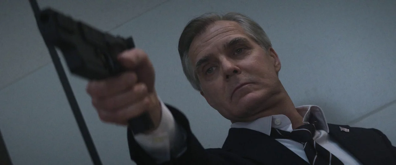 Mission: Impossible – Dead Reckoning Part One (2023) movie still: medium — A man in a suit stands in a hallway with plain white walls. He points a…