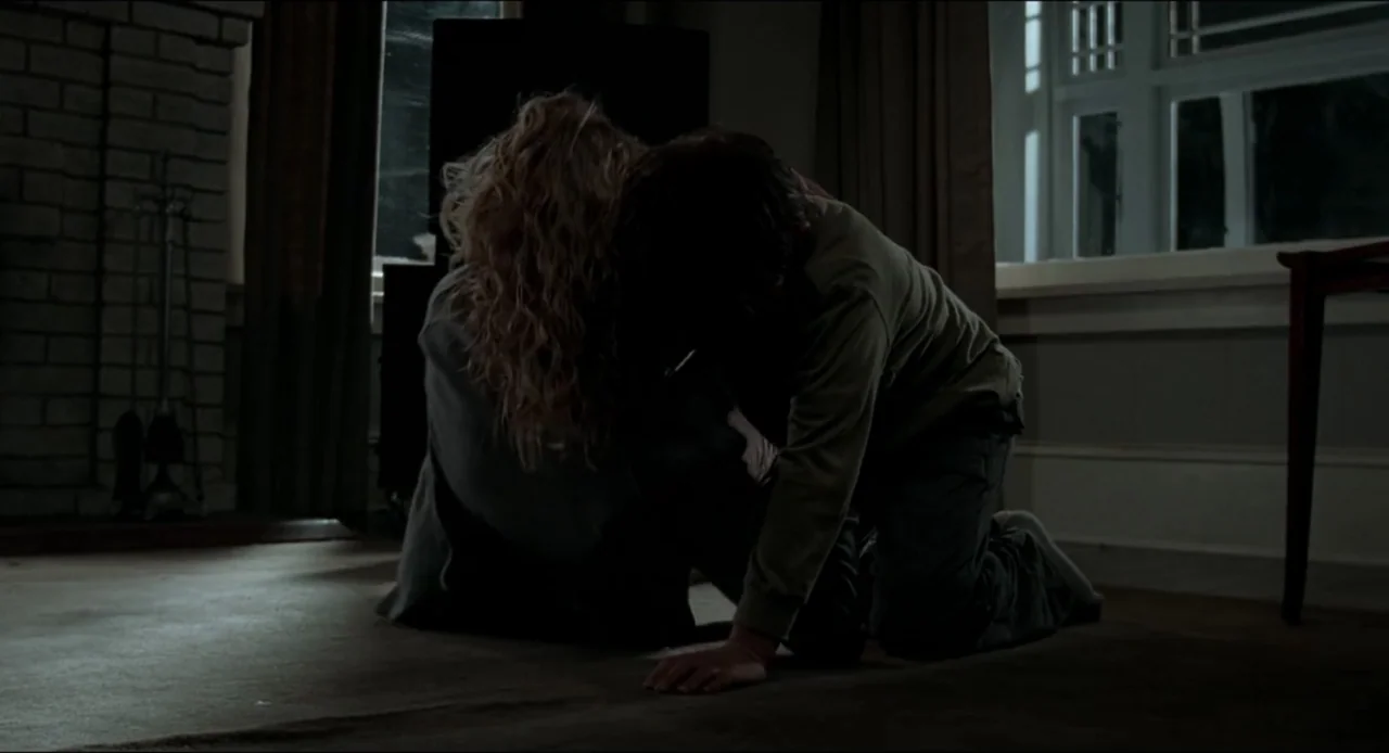 The Ring Two (2005) movie still: medium — A dimly lit room at night features a brick fireplace and a large window. Two people kneel on the carpet,…