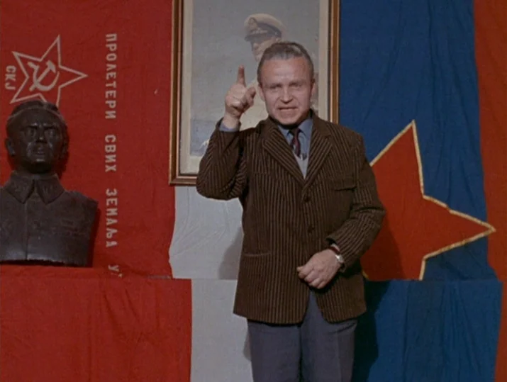 Innocence Unprotected (1968) movie still: medium — A man stands before a wall draped in communist red flags and a portrait. He gestures upward with…