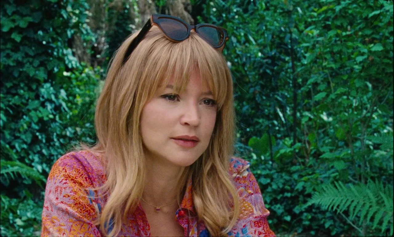 Just The Two Of Us (2023) movie still: medium — A woman sits outdoors surrounded by dense green vegetation including ferns and ivy vines. She stares…