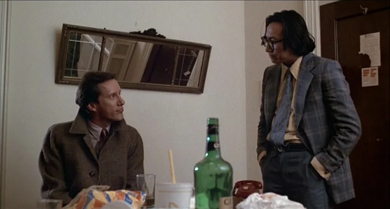 Videodrome (1983) movie still: medium — A hotel room with beige walls and a small rectangular mirror hanging above. Two men are interacting while…