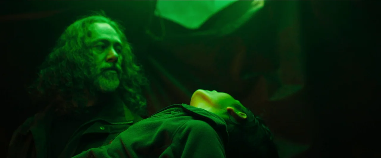 In The Earth (2021) movie still: medium — A dark, dimly lit environment is bathed in a saturated green light. A man with long hair carries a small,…