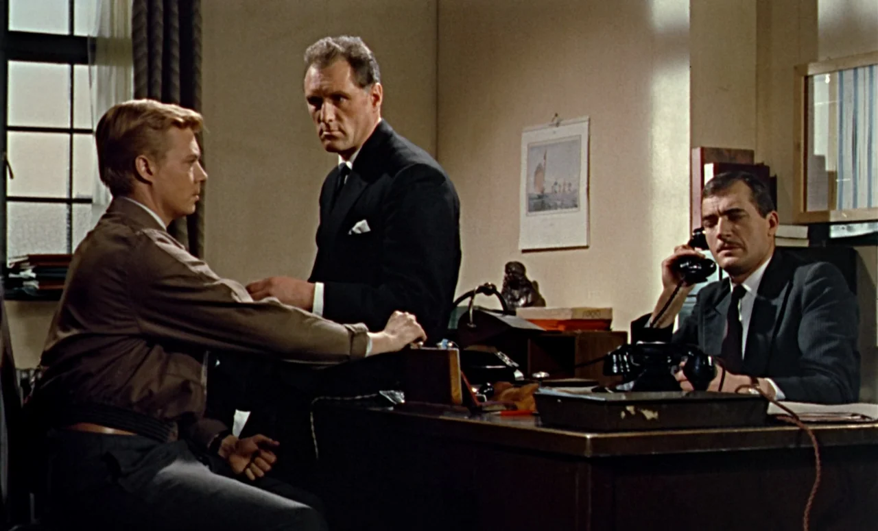 Peeping Tom (1960) movie still: medium — A mid-century office features beige walls, a wooden desk, and a window with curtains. Three men interact…