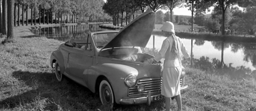 Les Amants (1958) movie still: wide — A quiet rural canal bank lined with tall, slender trees and lush green grass. A woman in a white suit stands…