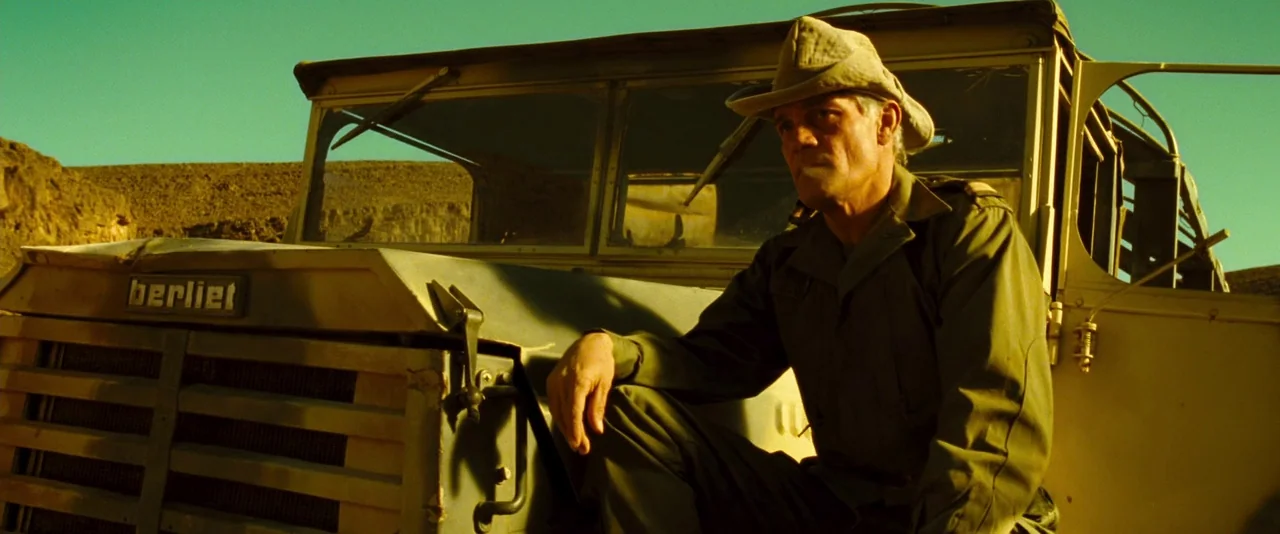 Micmacs (2009) movie still: medium — A rugged desert environment features a vintage olive drab truck under bright sun. A middle-aged man in military…
