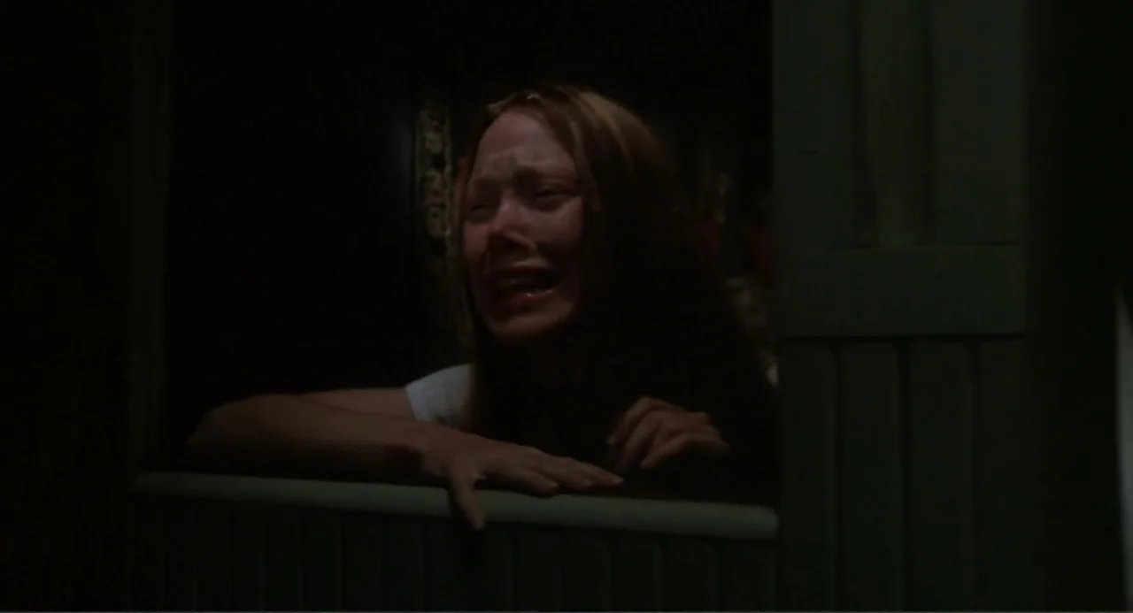 Carrie (1976) movie still: medium — A dark interior with a wood-paneled doorway frame partially obscuring the view. A young woman with long hair…