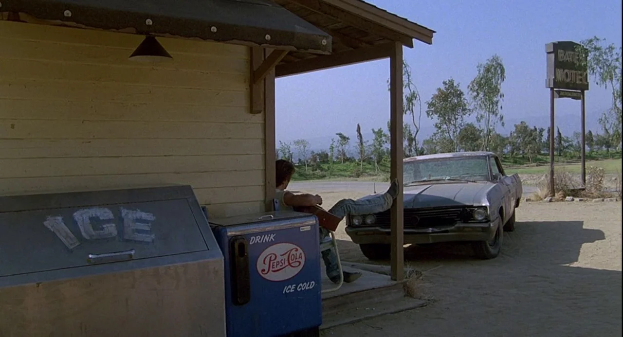 Psycho III (1986) movie still: wide — A rural motel entrance with a yellow exterior wall and a blue ice cooler. A man sits in a chair on the porch…