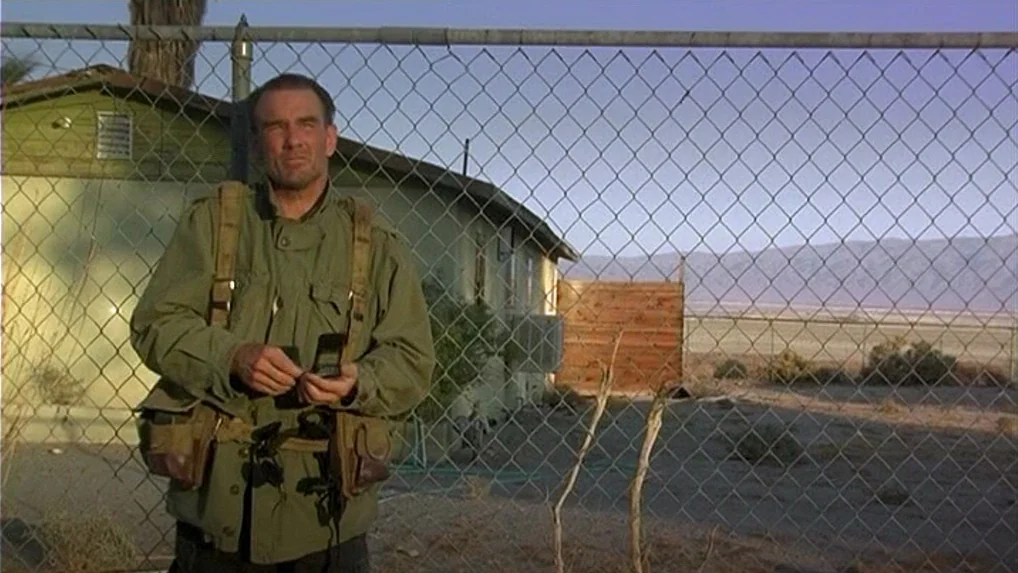 Land of Plenty (2004) movie still: medium — A man stands before a chain link fence in a dry, desert location. Behind him, a small structure and…