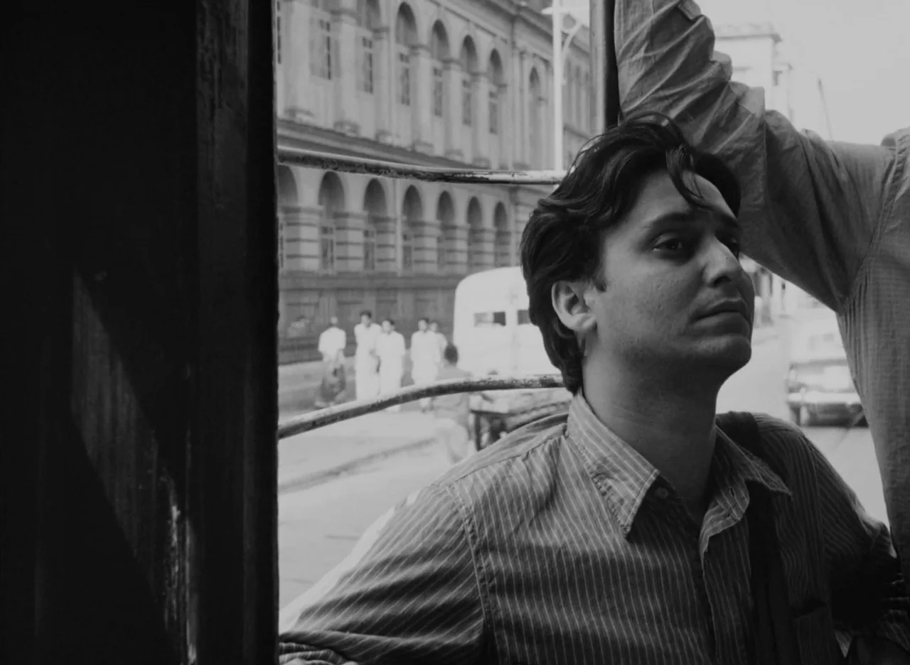 Apur Sansar (1959) movie still: medium — A man stands inside a vehicle looking through a barred window at a street. Behind him, stone buildings with…