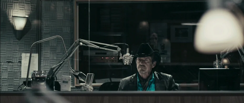 Pontypool (2008) movie still: medium — A dimly lit recording booth with acoustic wall tiles and visible glass partitions. An older man wearing a…