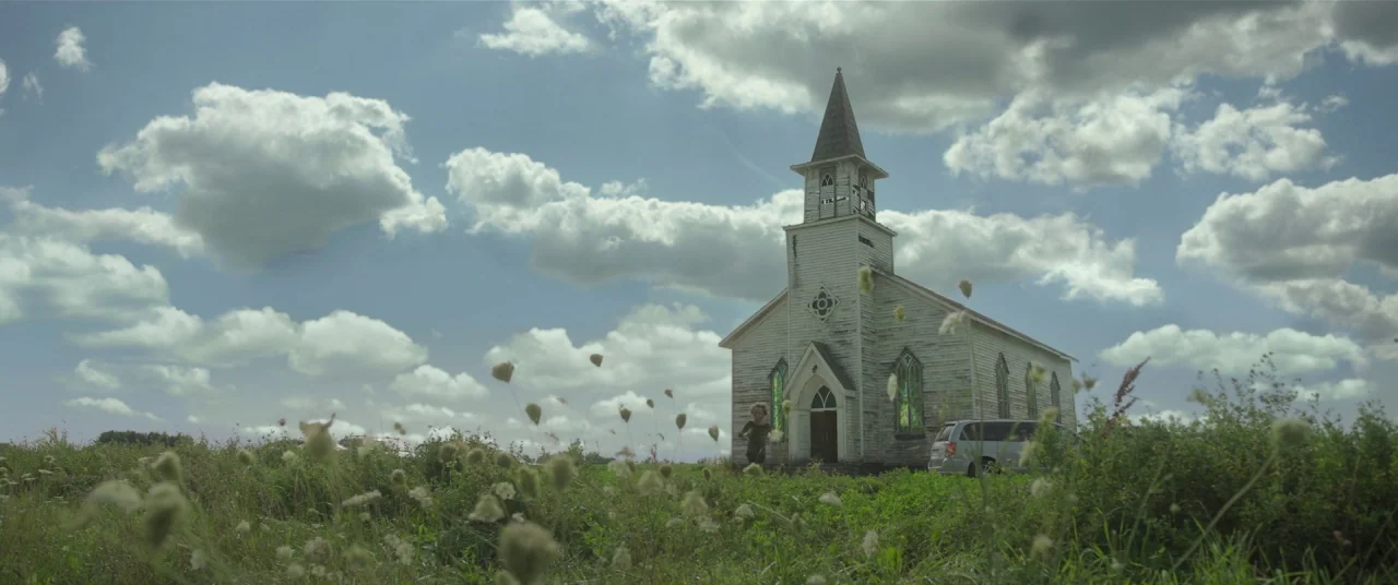 In The Tall Grass (2019) movie still: wide — A weathered white wooden church sits in a grassy field under a cloudy sky. A silver minivan is parked…