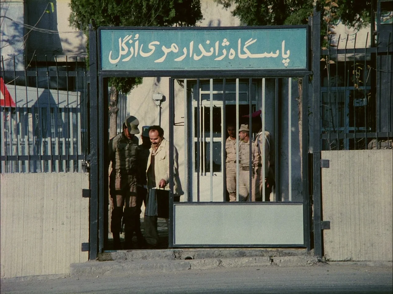 Close-Up (1990) movie still: medium — A gated entrance features a blue sign with Persian script mounted above vertical bars. Two uniformed men stand…
