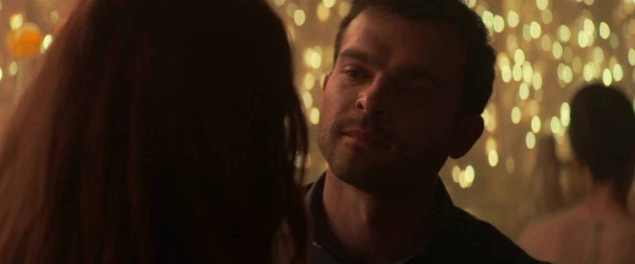 Fair Play (2023) movie still: closeup — A dimly lit room features warm out-of-focus light bokeh in the background. A man stands in the foreground…