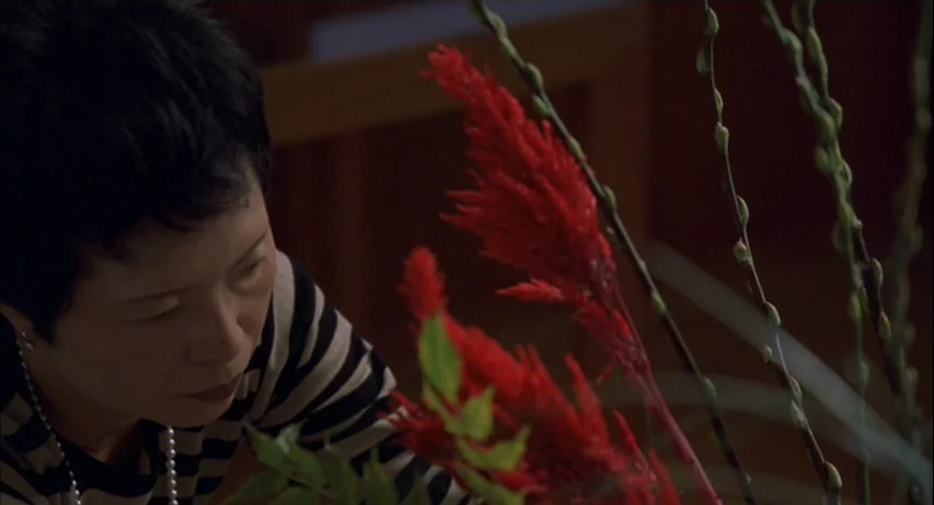 Lost In Translation (2003) movie still: closeup — A dimly lit indoor space contains a floral arrangement of red celosia. A woman with short dark…