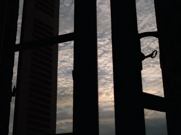 Keep Your Right Up (1987) movie still: establishing — A view through a darkened window frame shows a clouded daytime sky. Part of a building facade…