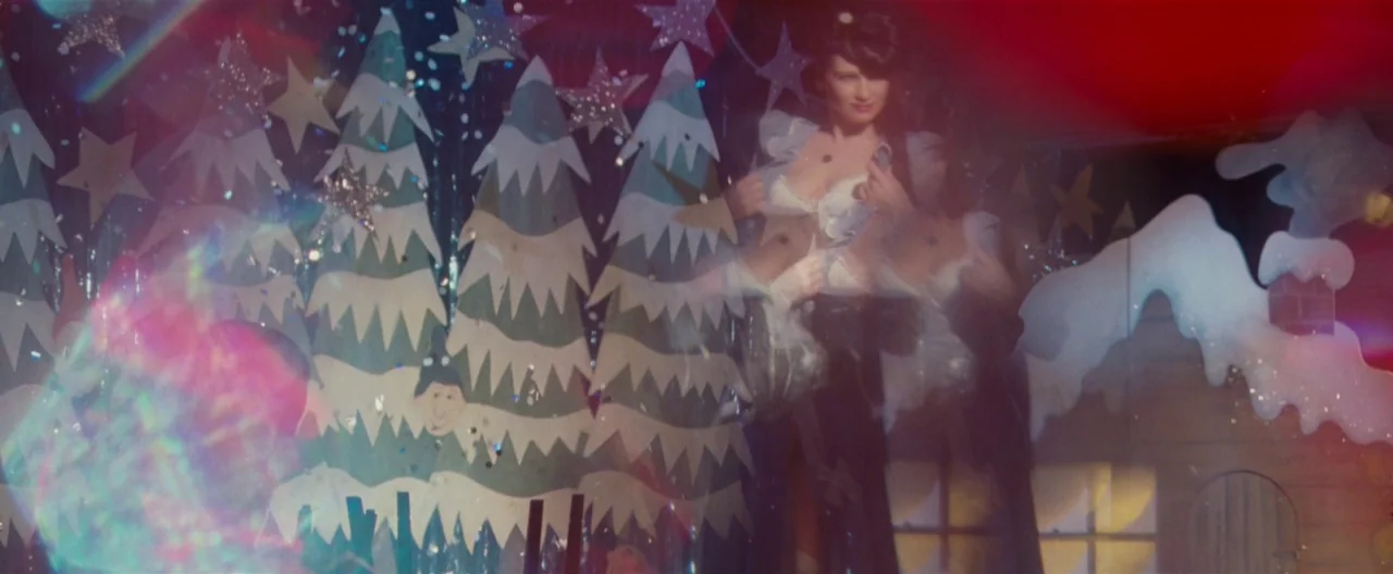 Hanna (2011) movie still: medium — A theatrical stage set features painted cardboard Christmas trees and a small log cabin. A woman in a festive…
