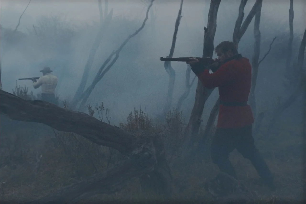 The Settlers (2023) movie still: wide — Two men stand in a sparse, dead woodland filled with thick grey fog. Each man holds a rifle aimed forward…