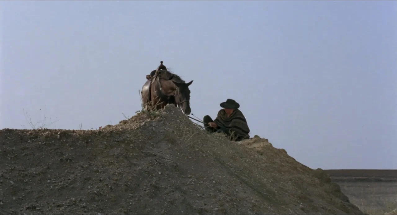 A Stranger in Town (1943) movie still: wide — An arid dirt mound stands against a clear, pale blue sky in a desert. A man in a poncho and hat sits…