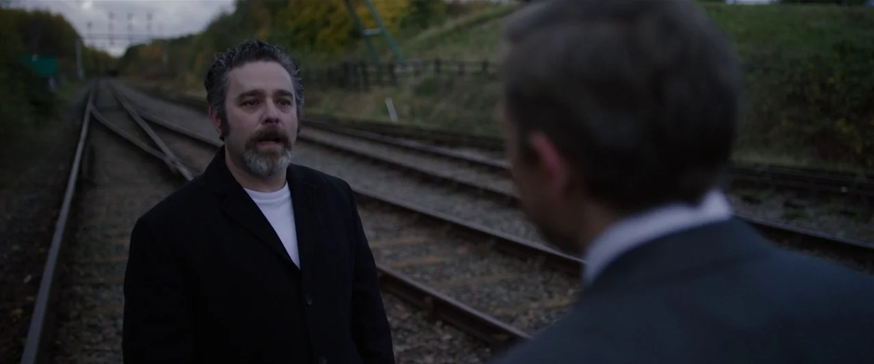 Ghost Stories (2017) movie still: medium — A pair of parallel railway tracks stretches into the distance through a grassy area. A middle-aged man…