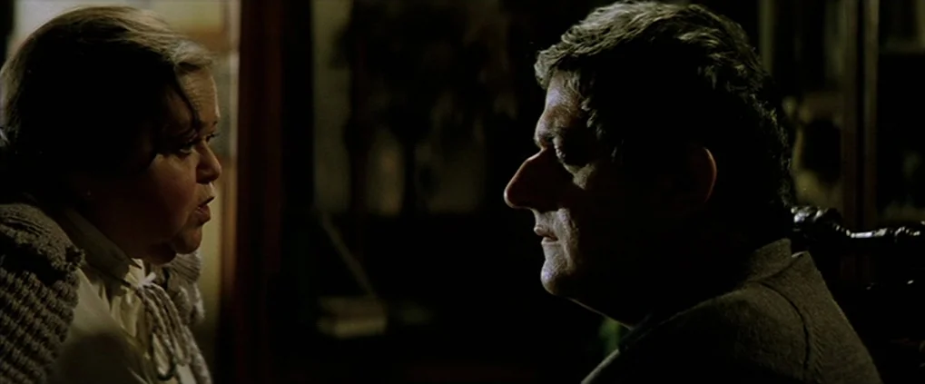 Anguish (1987) movie still: medium — A dark indoor space contains an older man and woman facing each other. They sit in a dimly lit room with muted…