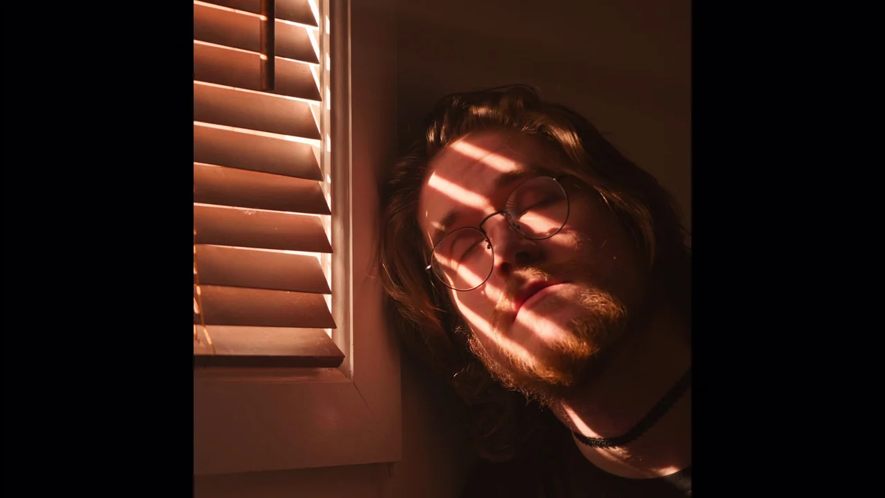 Bo Burnham: Inside (2021) movie still: closeup — A man stands indoors next to a white window frame with slatted blinds. He leans his head against…