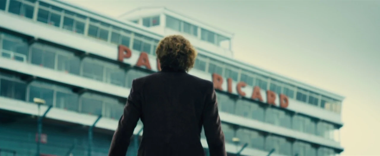 Rush (2013) movie still: fullbody — A modern multi-story grandstand building looms under a clear sky. A lone person stands in the foreground facing…