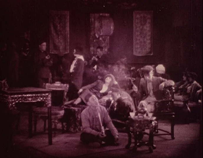 Broken Blossoms (1919) movie still: wide — A dimly lit room filled with hazy smoke and hanging rectangular tapestries on walls. Multiple figures are…