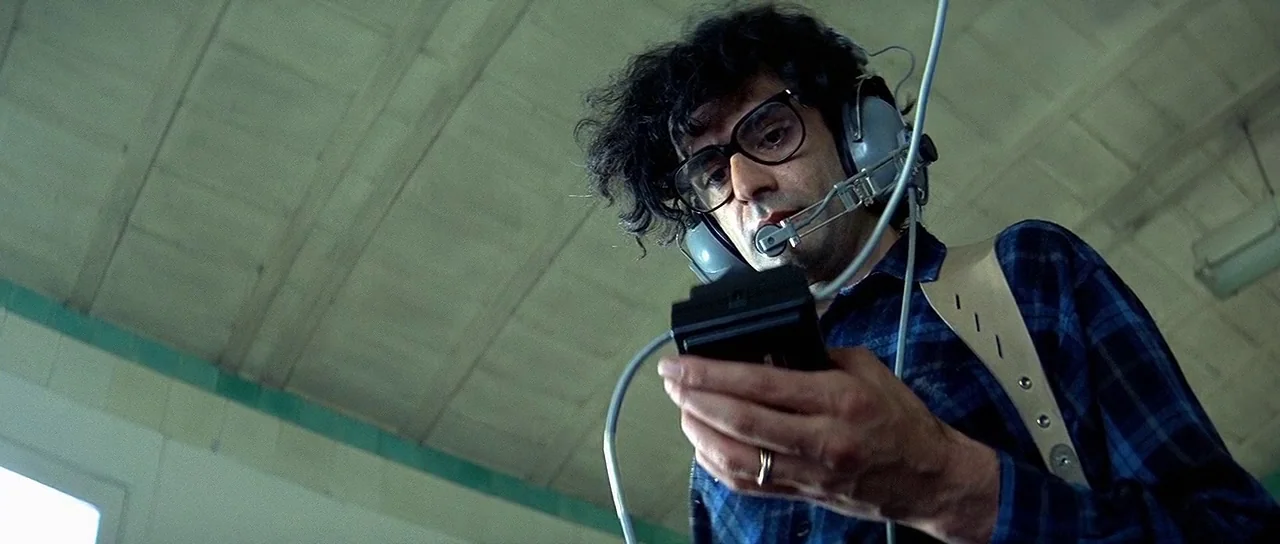 Nikita (1990) movie still: medium — An industrial space with a light green ceiling and exposed structural beams is visible. A man wearing headphones…