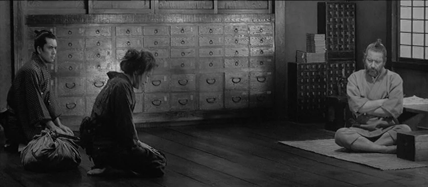 Red Beard (1965) movie still: wide — An apothecary interior features rows of wooden apothecary drawers lining the back wall. Two visitors kneel on…