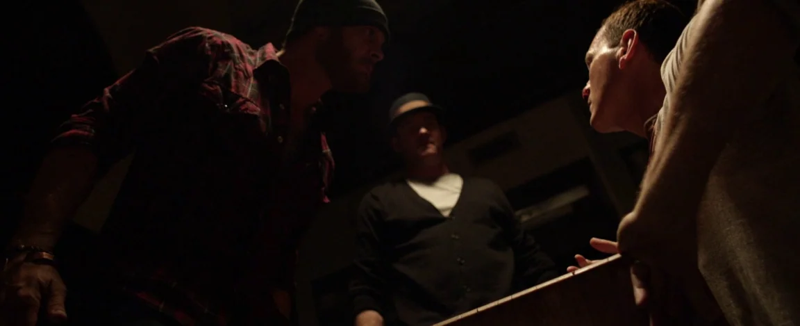 Cheap Thrills (2013) movie still: medium — Three men stand around a dark, shadowed table in a low-ceilinged interior room. They are positioned in a…
