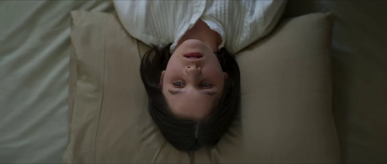 The Sacrifice Game (2023) movie still: closeup — A soft beige pillow rests beneath the head of a person against a sheet. The person lies on their…