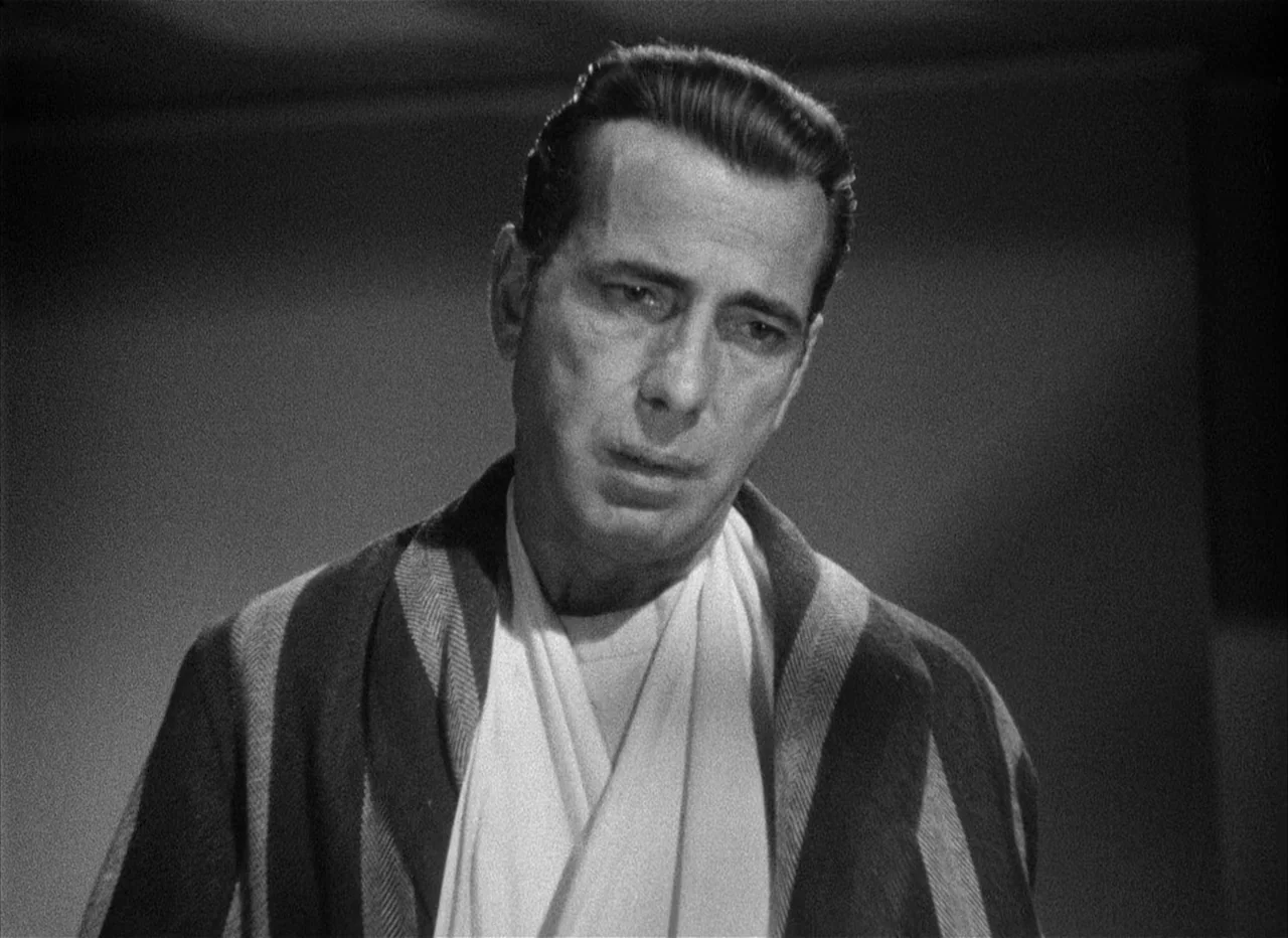 Dead Reckoning (1946) movie still: medium — A man in a house robe stands against a dark, plain background. The focus is entirely on his face as he…