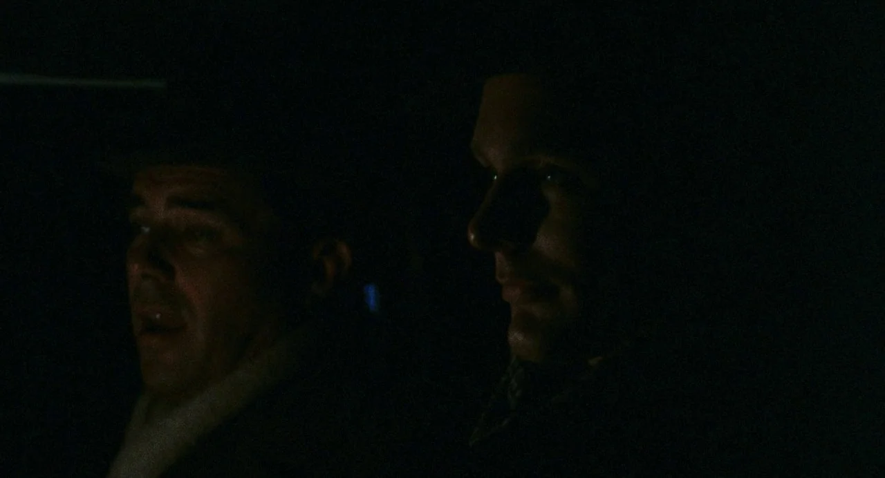 The Damned (1969) movie still: medium — The interior of a vehicle at night is illuminated by passing external streetlights. Two men occupy the front…