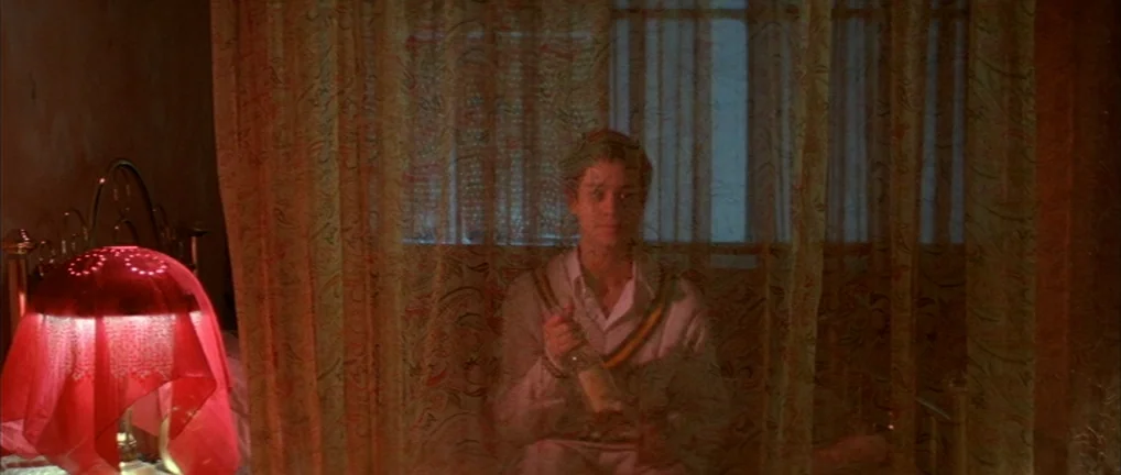 The Crying Game (1992) movie still: medium — A person sits behind sheer patterned curtains in a dim, red-lit bedroom. A bedside lamp with a…