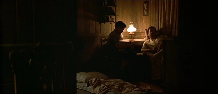 Road To Perdition (2002) movie still: wide — A dimly lit room with wooden walls and a small table lamp. A boy sits at a desk while a man lies on a…
