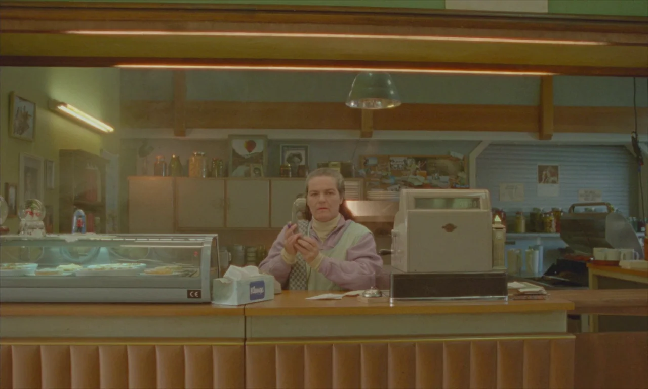 Spencer (2021) movie still: medium — A diner counter interior features a glass display case and vintage cash register. A woman stands behind the…