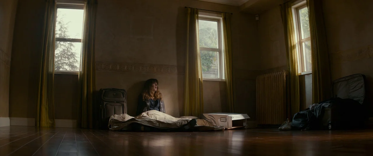 Colossal (2016) movie still: wide — An empty room with hardwood floors has windows draped in yellow curtains. A woman sits on the floor wrapped in a…