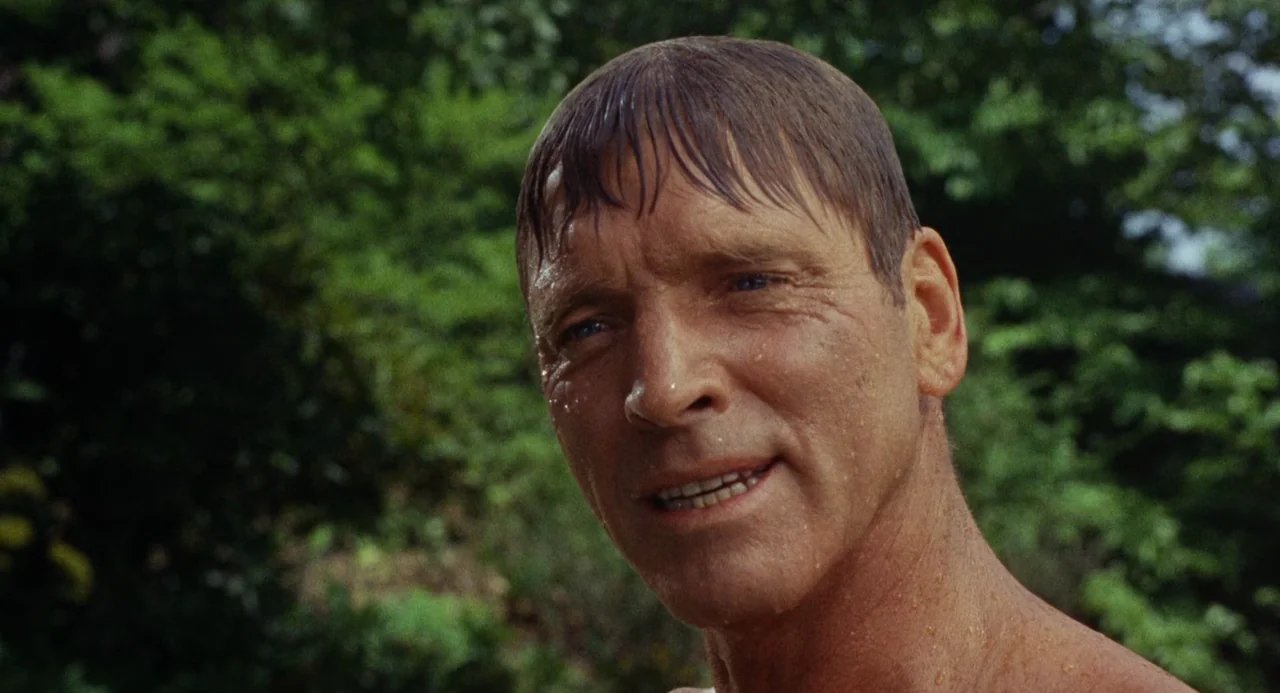 The Swimmer (1968) movie still: closeup — An outdoor area with dense blurred green foliage in the background. A man with wet skin and hair stands…